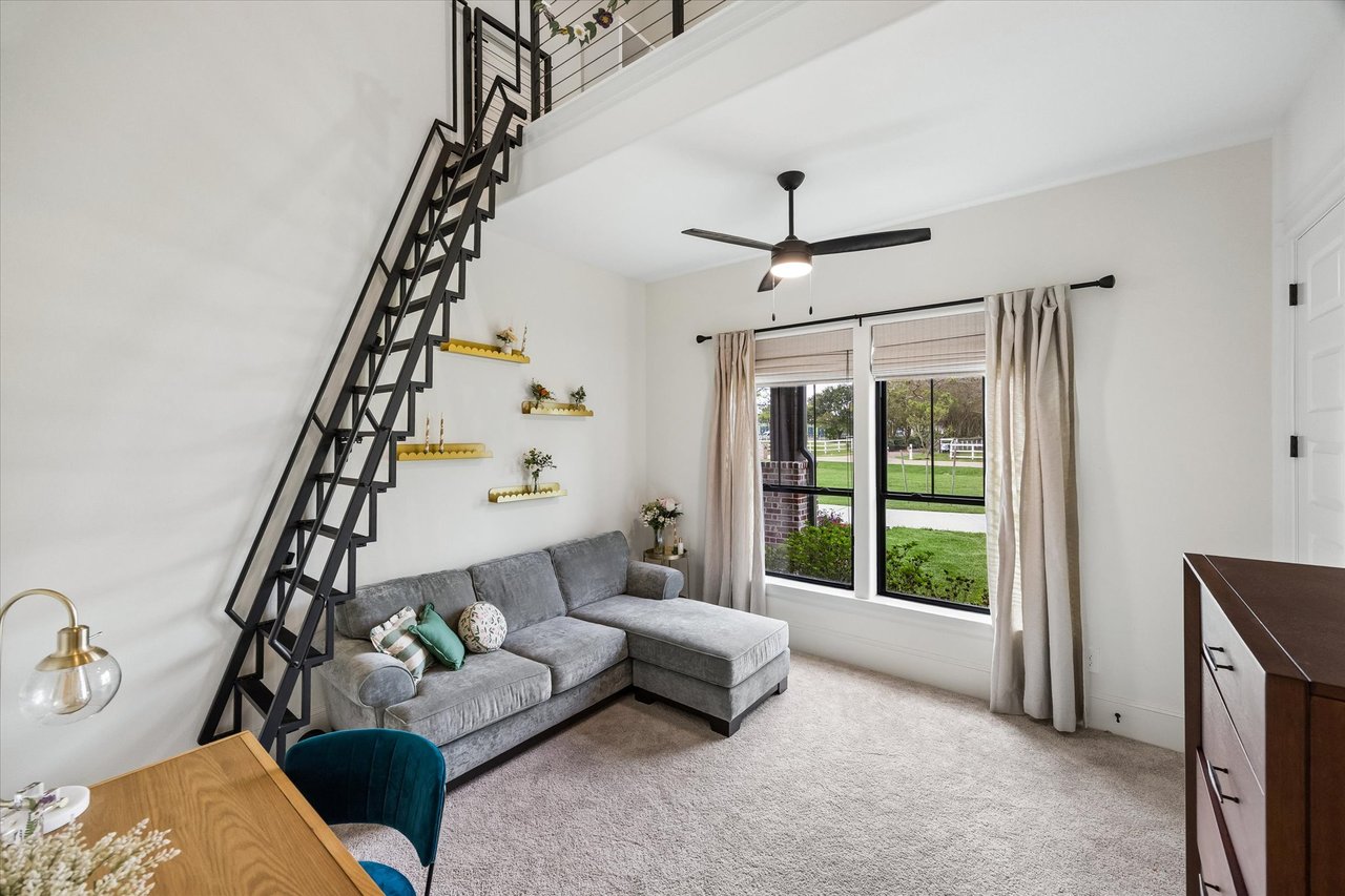 Living room with a gray sectional beneath a black staircase and windows overlooking a lawn.