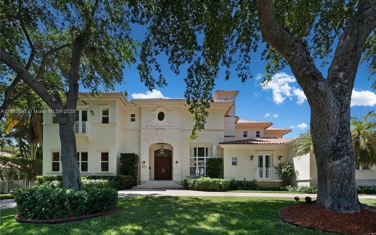 Cream-colored two-story home with an arched entry framed by mature trees and a landscaped lawn.