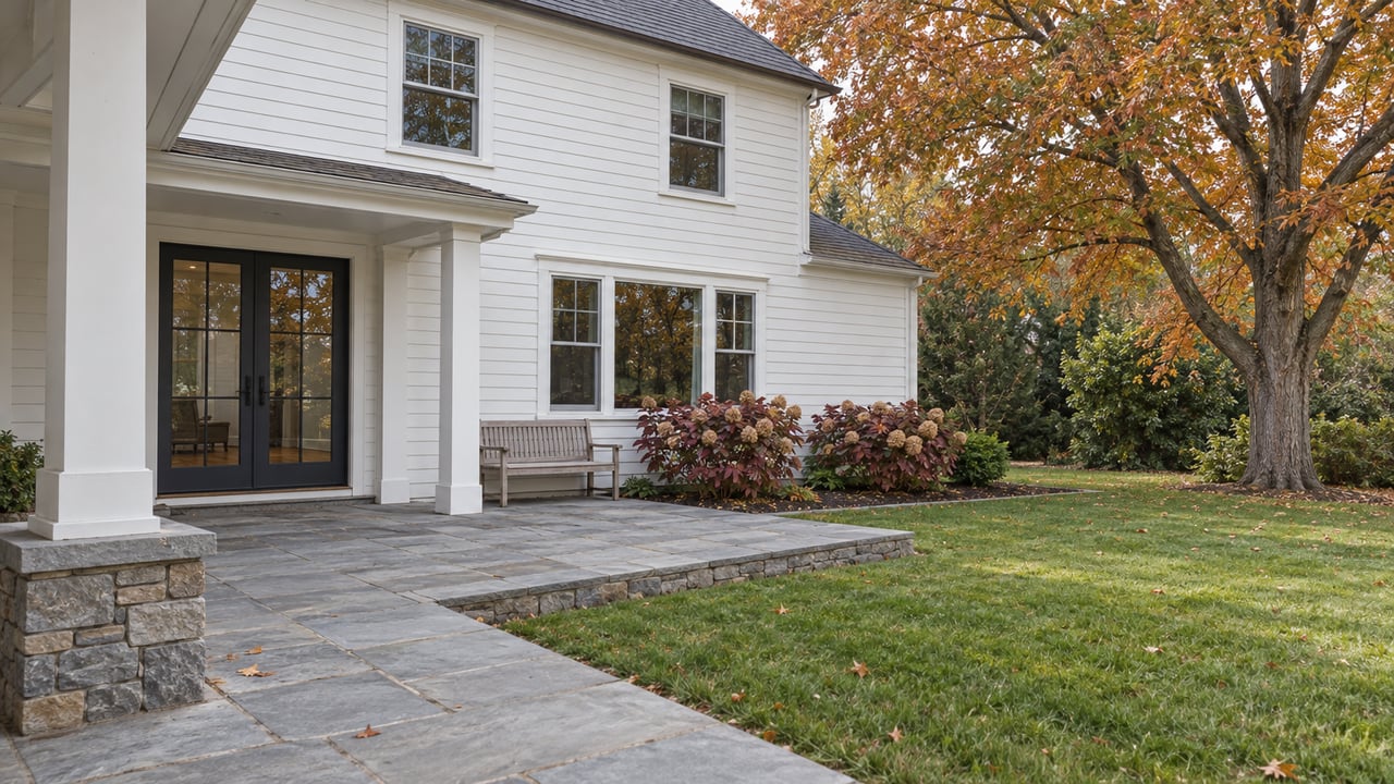 White clapboard home with a covered porch, bluestone terrace and autumn maple beside a spacious lawn.