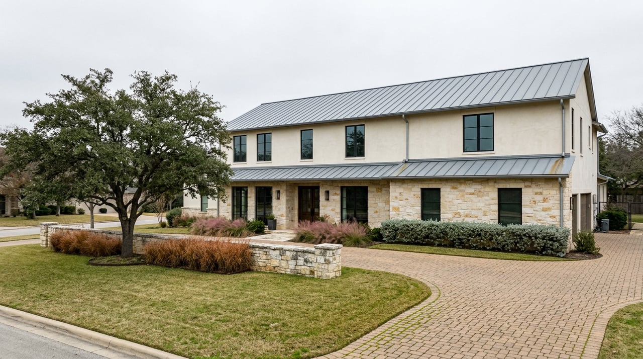 Two-story limestone-and-stucco home with black-framed windows, broad oak, lawn, and sweeping clay-paver motor court.