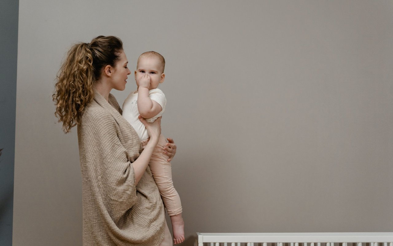 A woman holds a baby beside a white crib in a neutral-toned room.