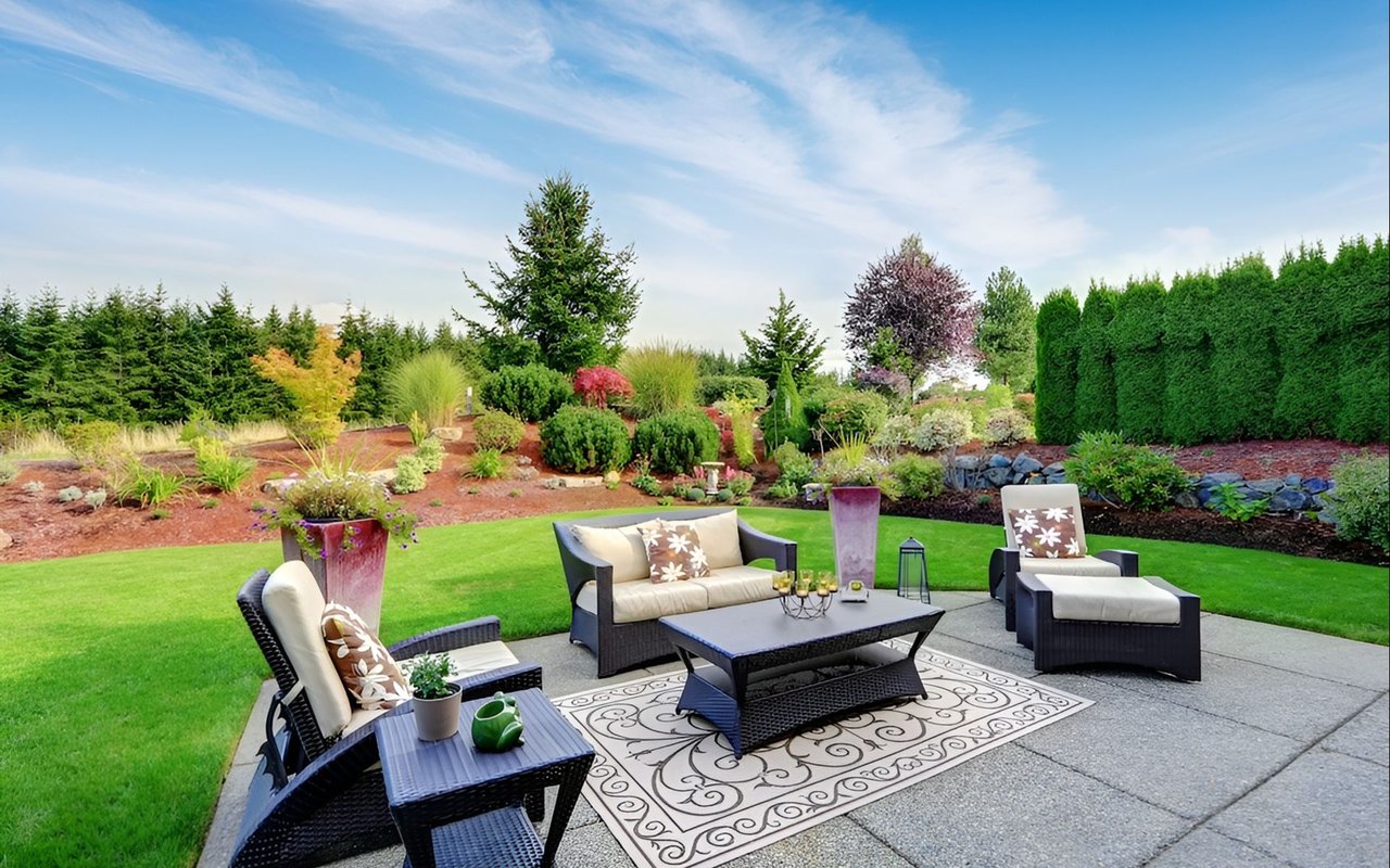 Furnished backyard patio overlooking a green lawn and layered landscaping with colorful shrubs and tall evergreen hedges.