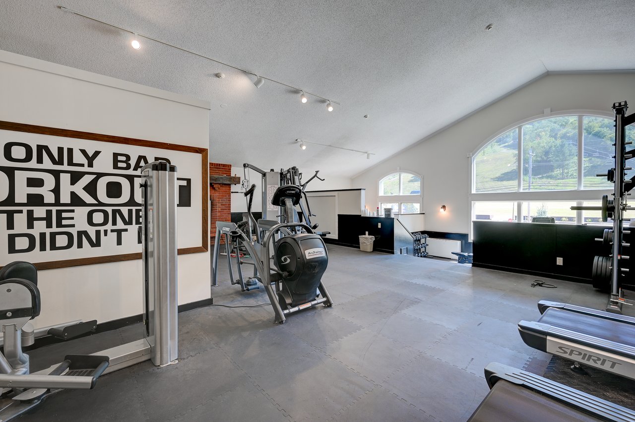 Condominium fitness room with cardio machines, weight equipment, and arched windows overlooking a green hillside.