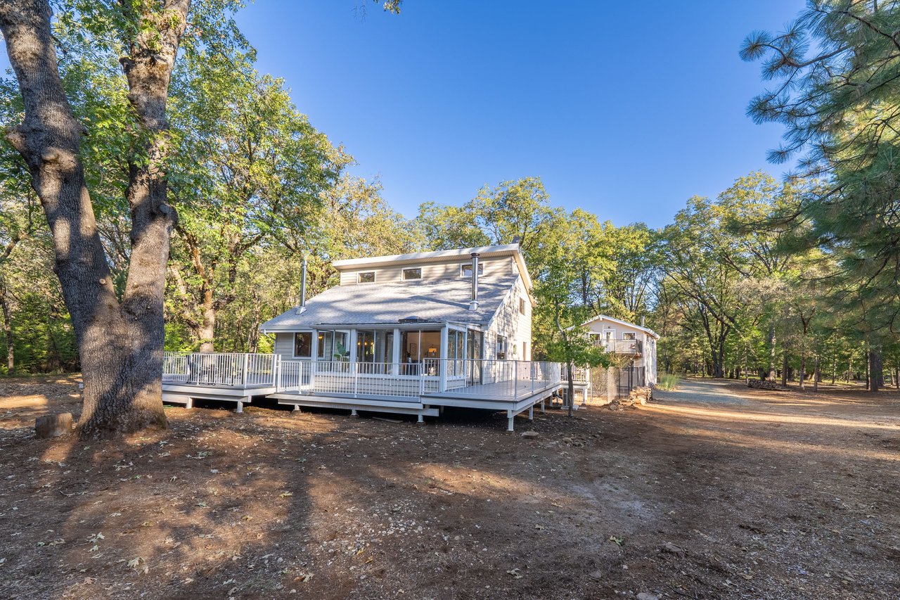 Two-story home with a wide wraparound deck among mature trees, with a small outbuilding nearby.