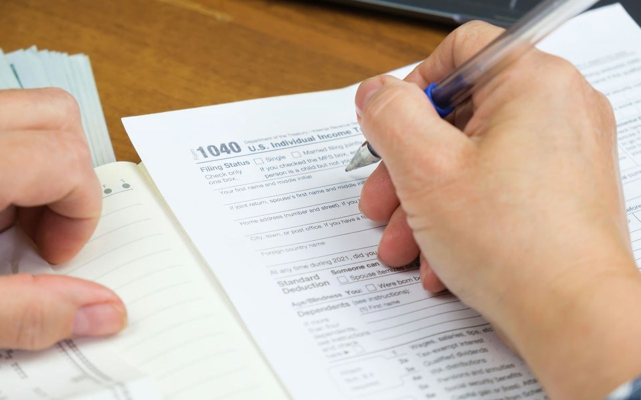 Hands filling out a 1040 income tax return beside an open notebook.