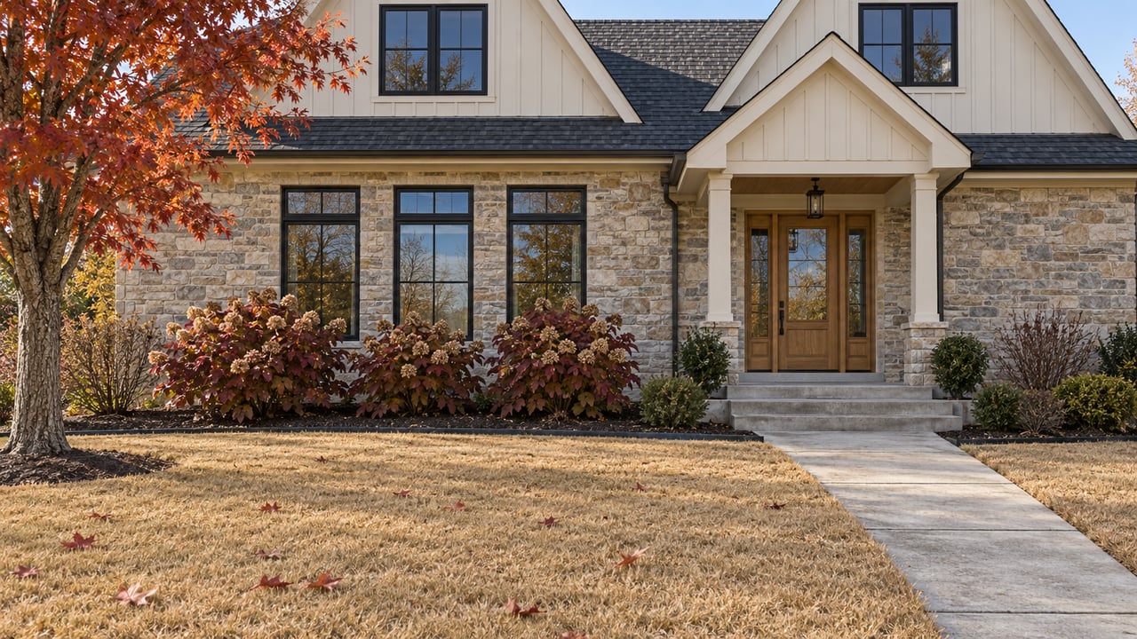 An ivory-sided, stone-clad home beyond a broad tawny lawn, with an oak entry door and a red maple in autumn color.