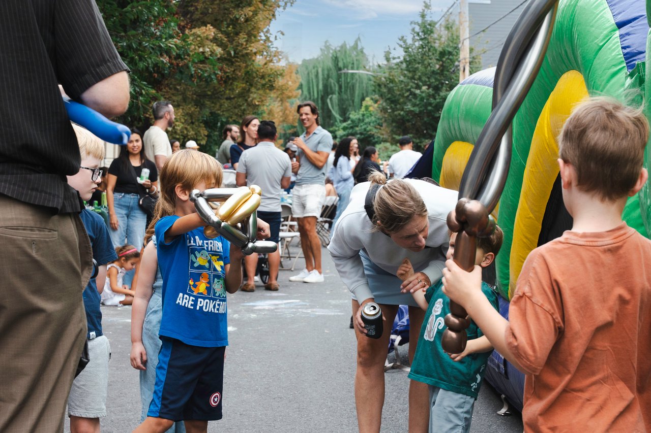 Neighbors mingle at a block party as children hold balloon animals beside a colorful inflatable slide.
