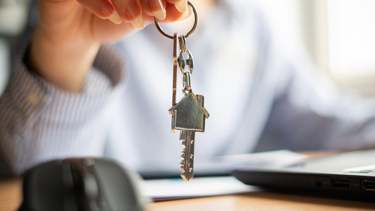 A house-shaped key pendant hangs from a key ring held above a desk.