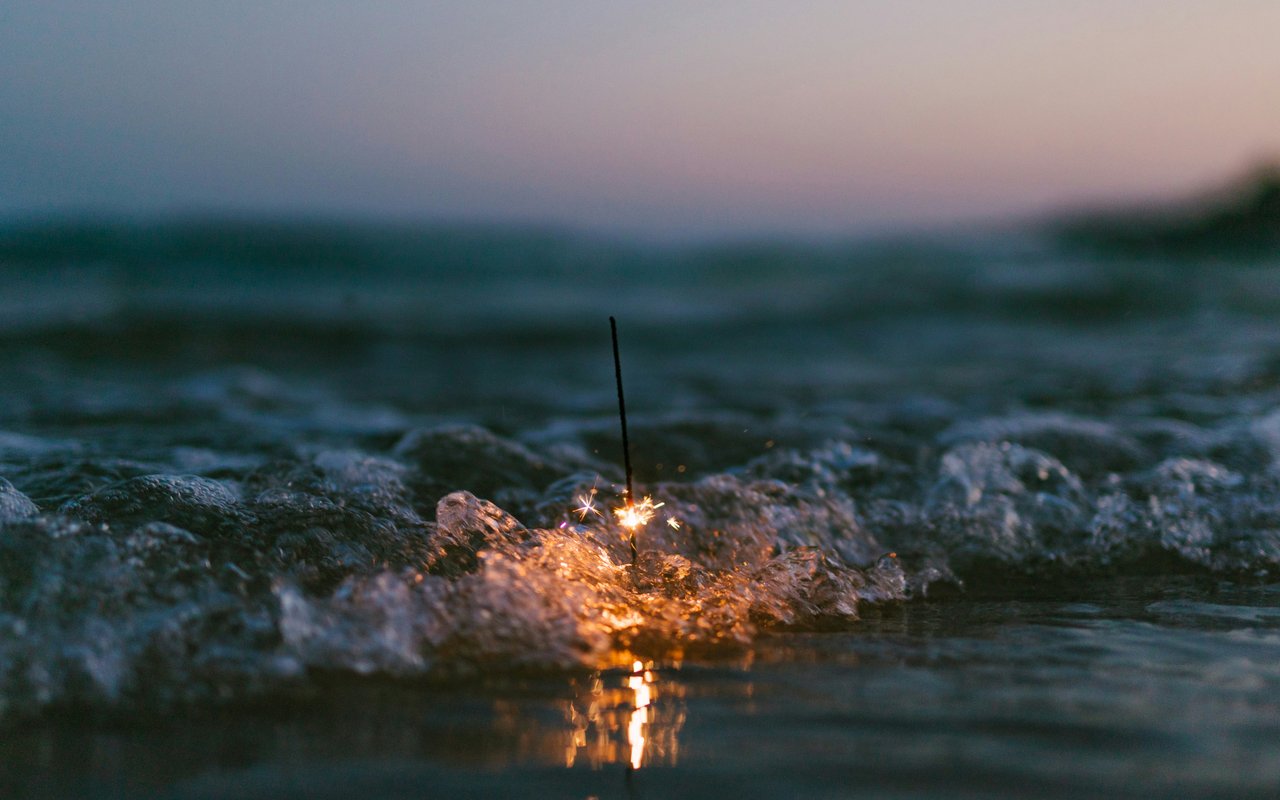 A close-up, low-angle shot of a sparkler burning in the water as a gentle wave washes over it at dusk.
