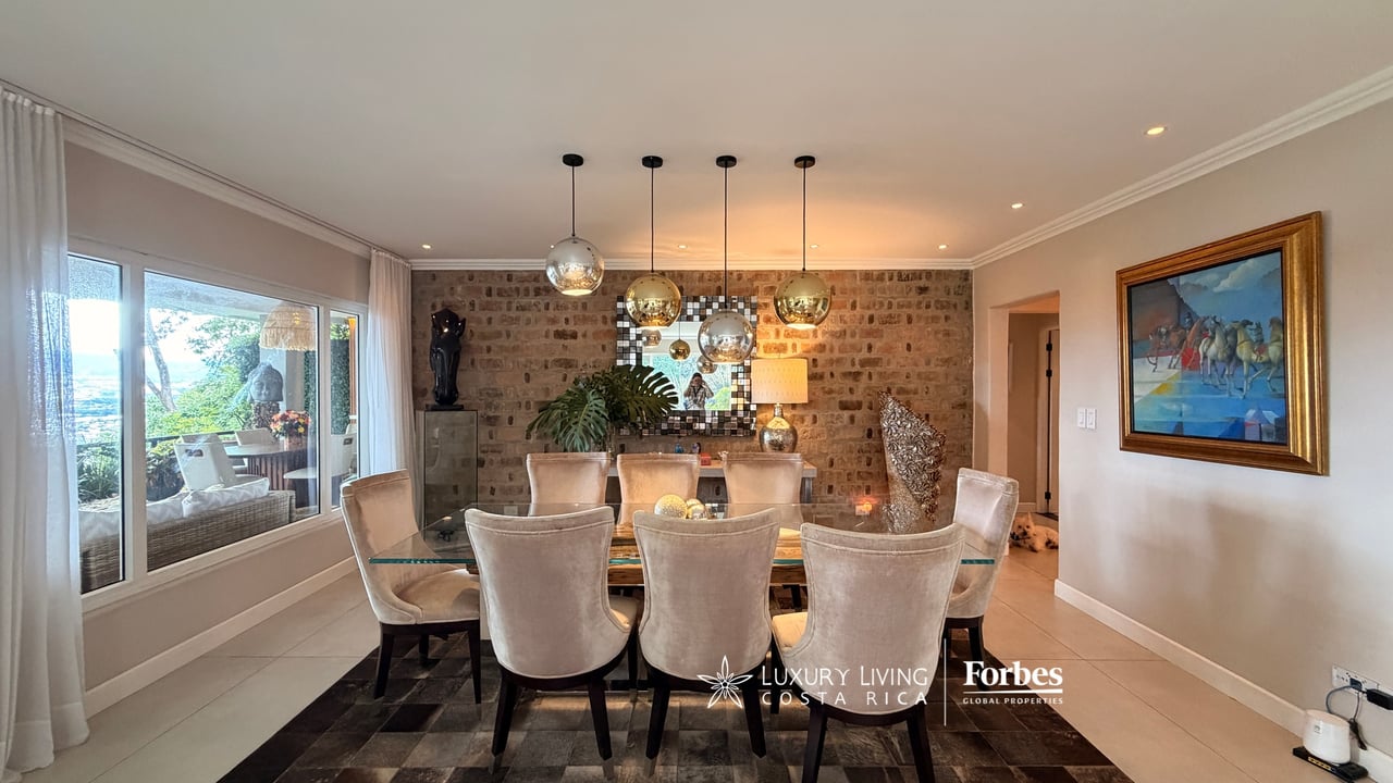 Dining room with a glass table, upholstered chairs, brick accent wall, and large garden-facing windows.