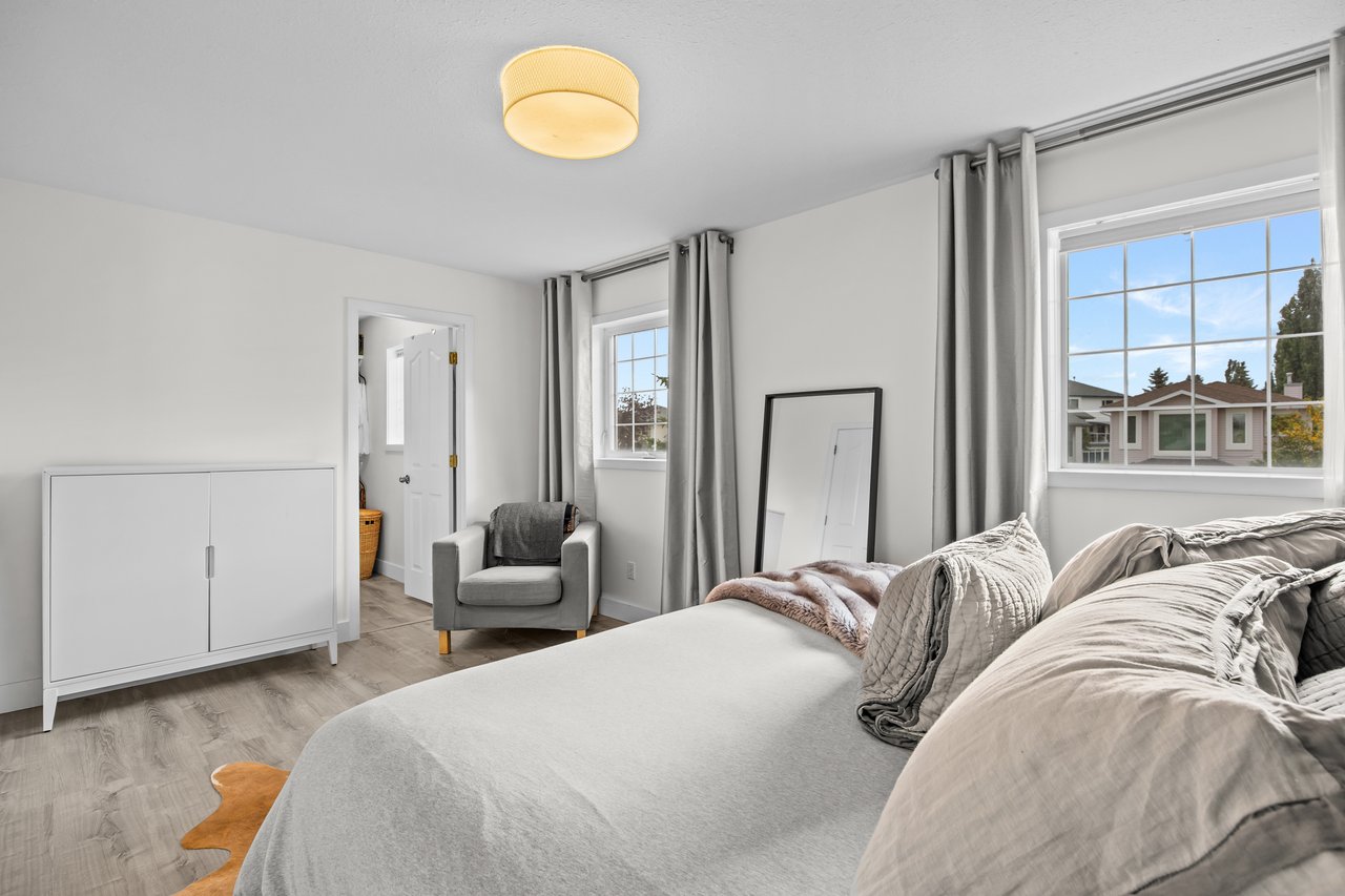 Bright bedroom with pale bedding, a gray armchair, and two curtained windows.