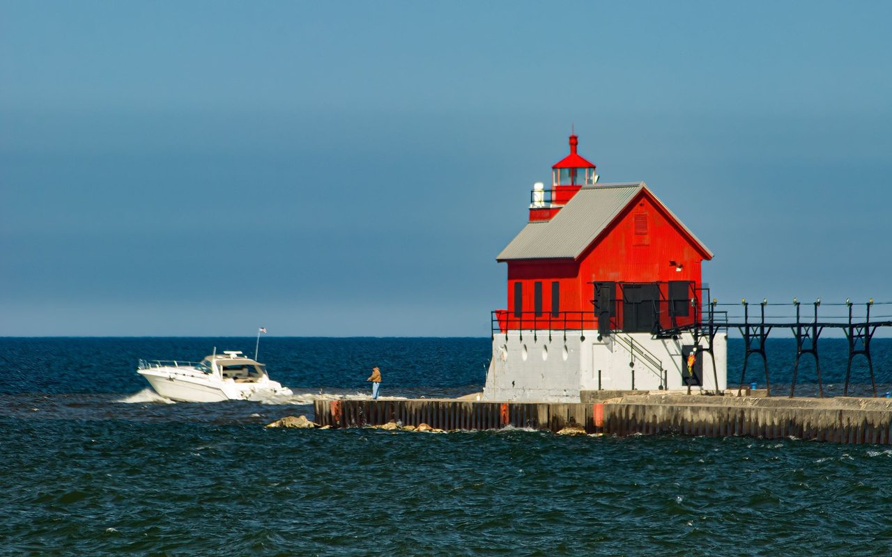 Red lighthouse on a pier over blue water, with a passing motorboat and a person on the pier.