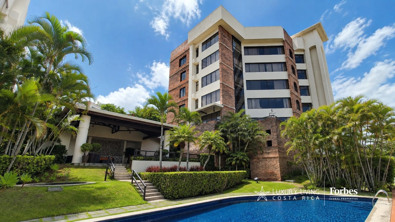 Escazú condominium with brick and white façades, palm trees, landscaped grounds, and a swimming pool.