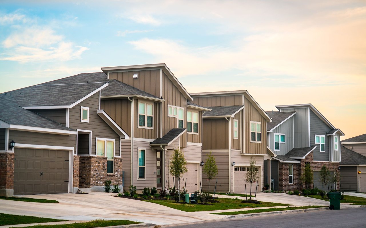 Attached homes with brown and gray siding, brick accents, and wide concrete driveways at sunset.