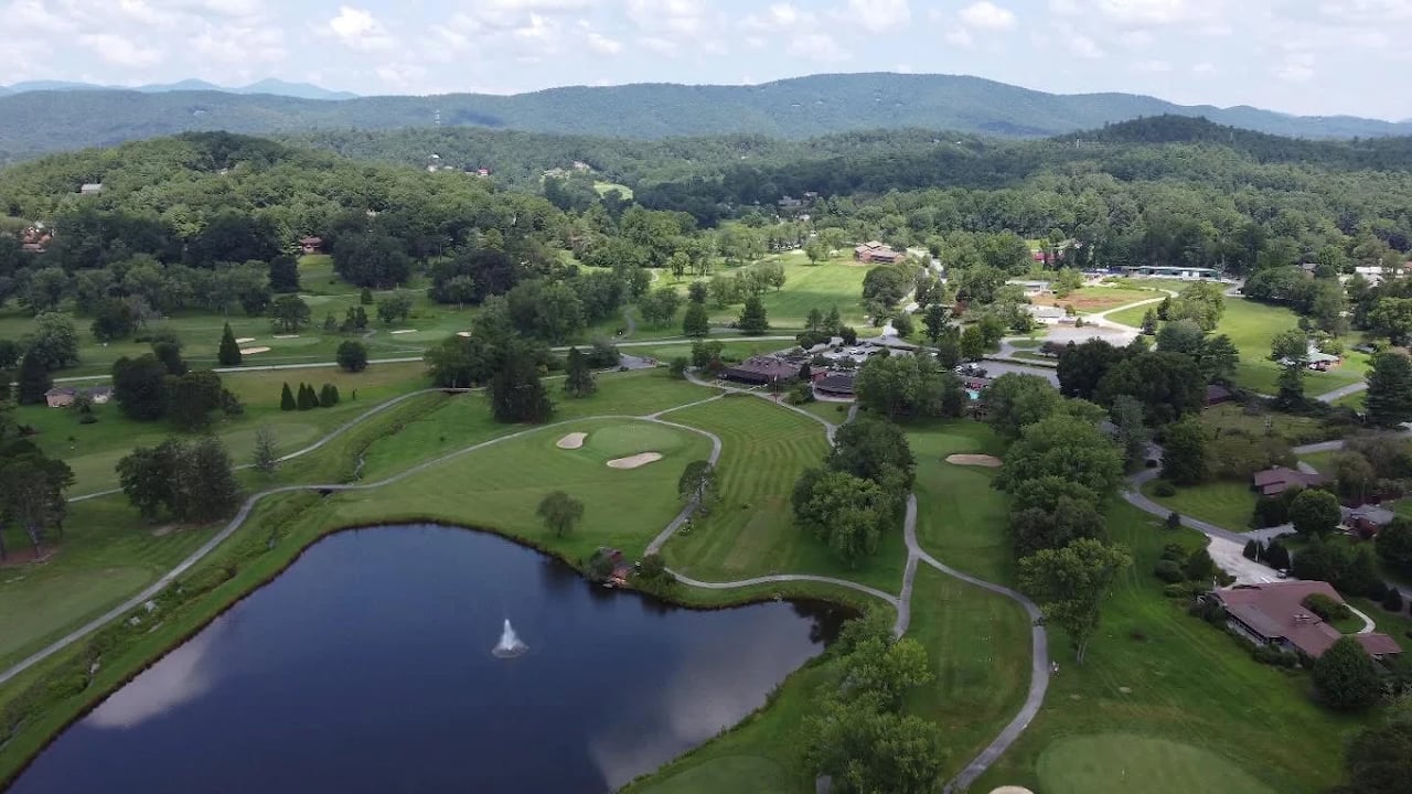 Aerial view of a golf course with a fountain pond, homes, and wooded mountains beyond.