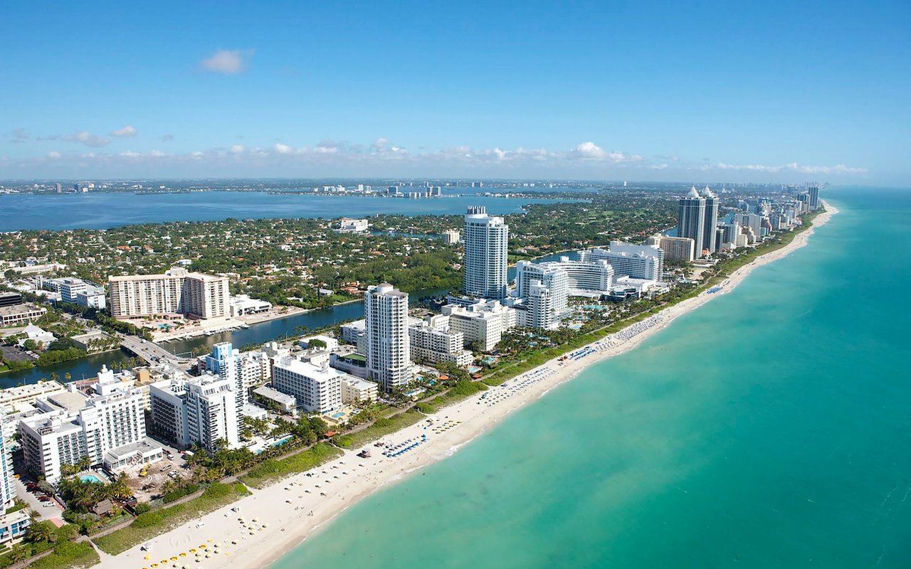 Aerial view of Miami Beach’s high-rises lining the sandy coast beside turquoise ocean water.
