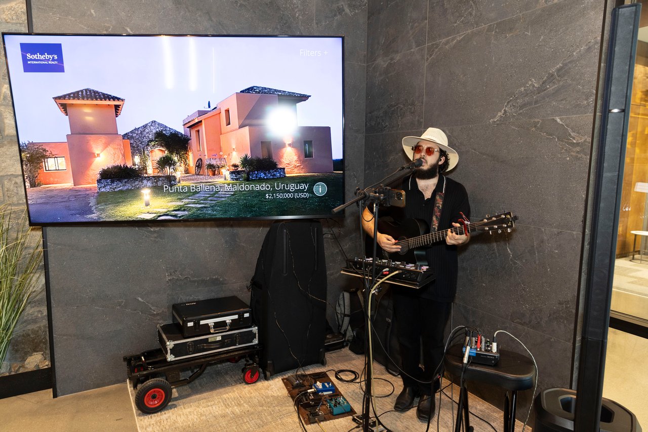 A guitarist performs beside a screen displaying a Sotheby's property listing.