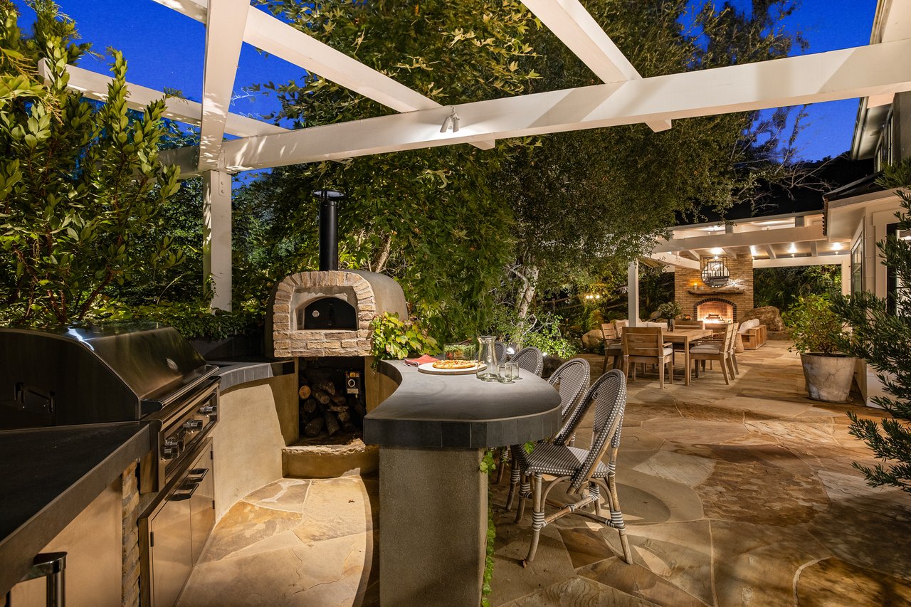 Outdoor kitchen with a stone pizza oven and pergola-covered dining area beside a lit fireplace.