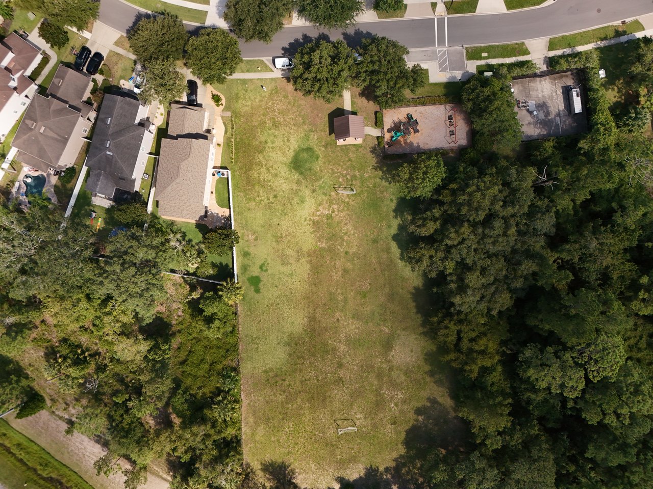 Aerial view of a broad grassy lot bordered by trees, neighboring homes, and a small playground.