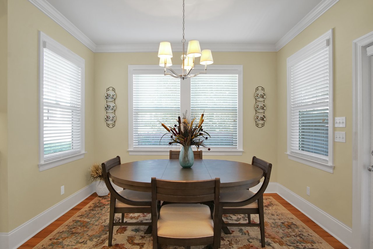 A cozy dining room with a round wooden table set for four. A vase with flowers sits at the center. Soft light filters through blinds on two windows.