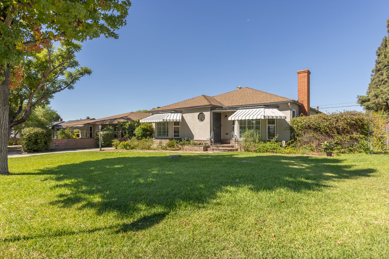 Single-story stucco home with striped awnings and a brick chimney beyond a broad green lawn.