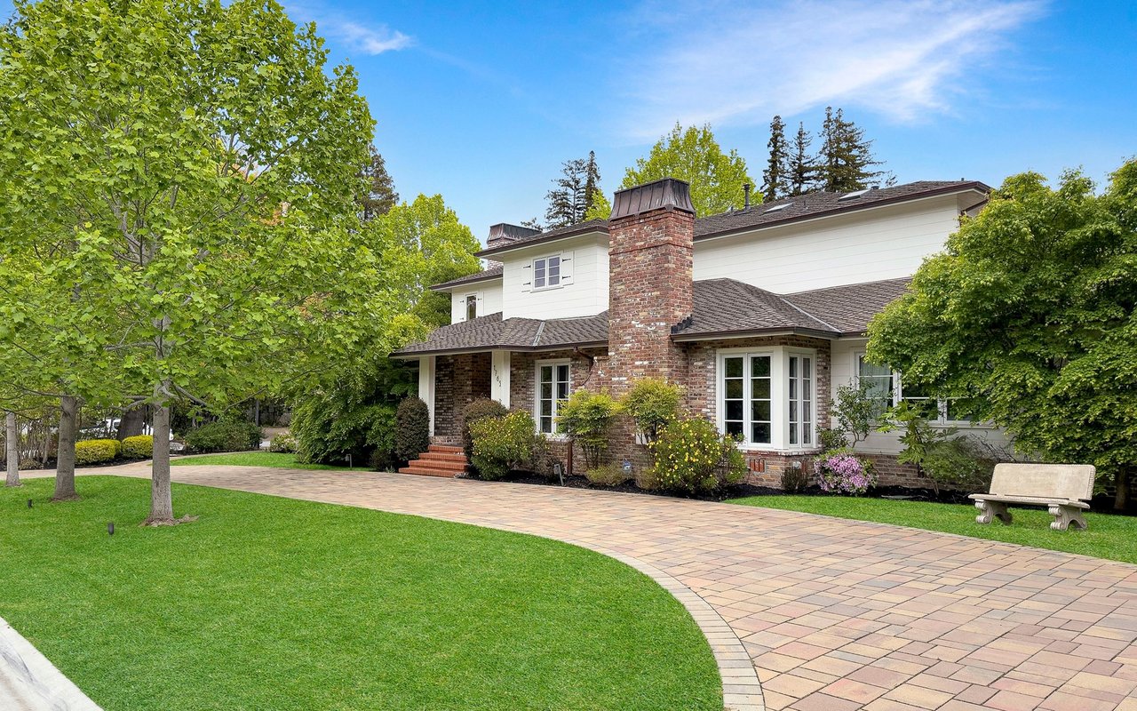 White two-story house with a brick chimney, curved paved driveway, manicured lawn, and mature trees.