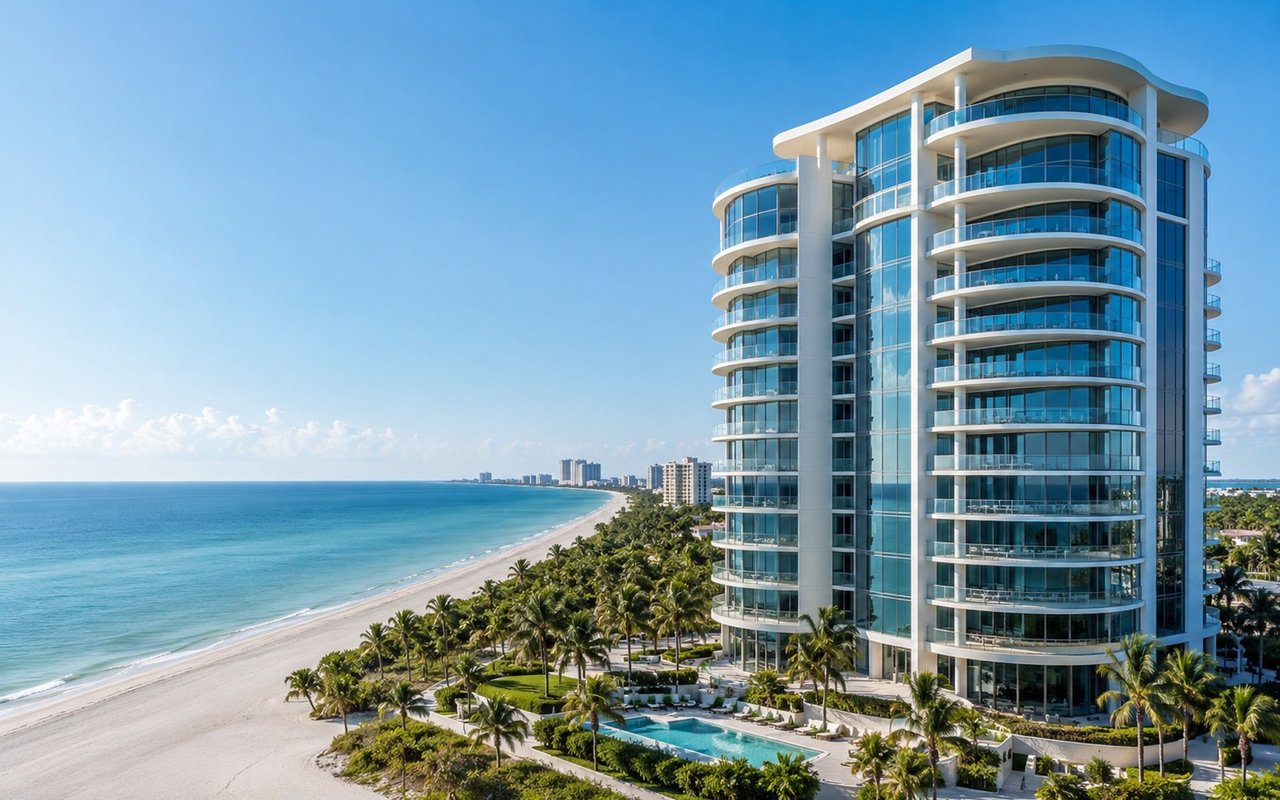 Oceanfront condominium tower with curved glass balconies beside a palm-lined beach and pool.