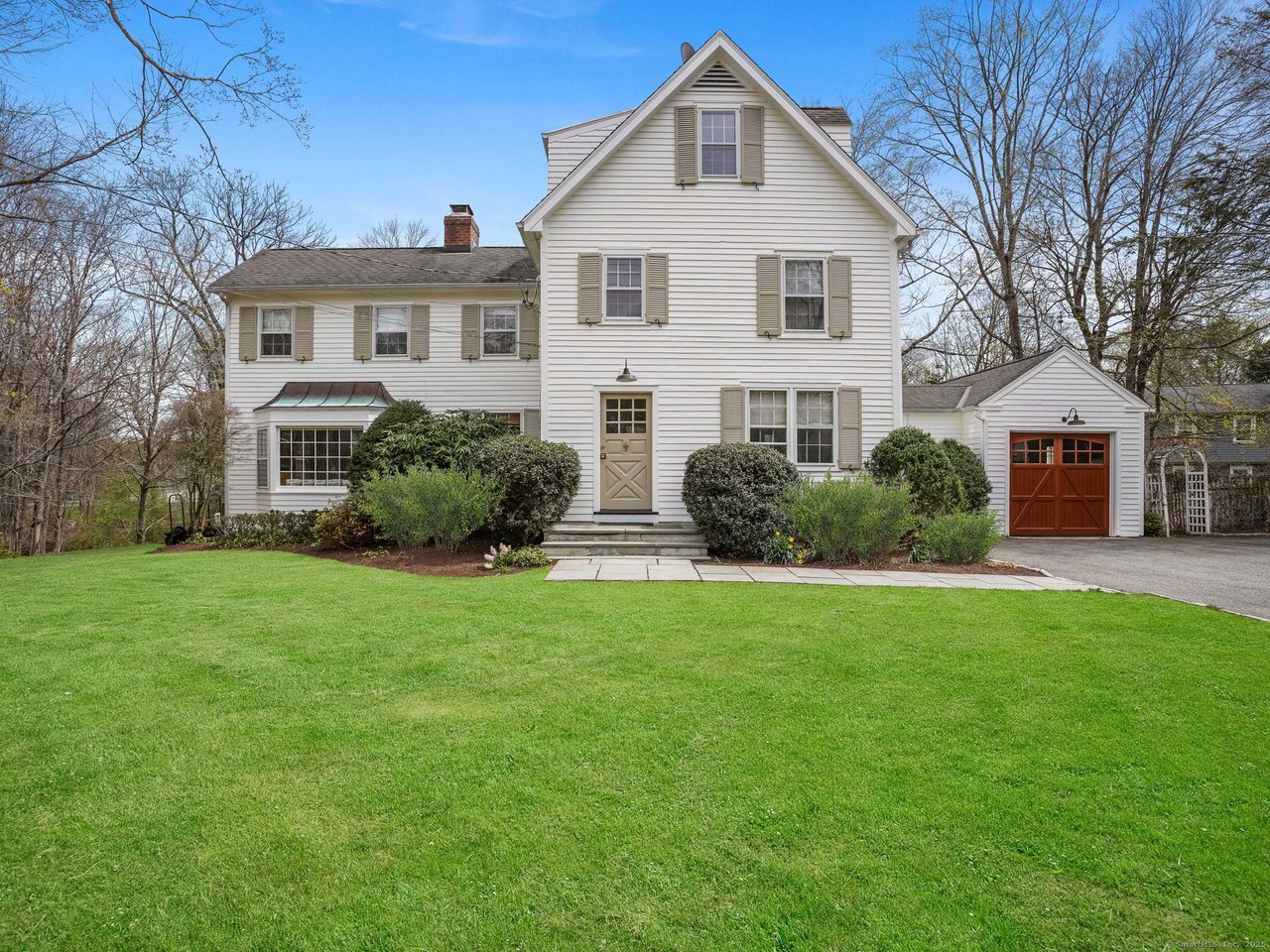 White two-story clapboard house with a steep front gable, red-brown garage door, and broad green lawn.