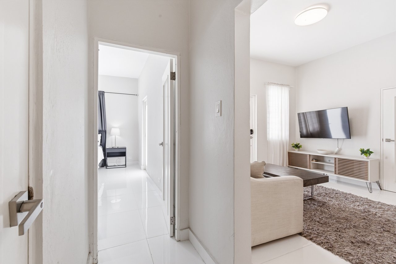 Furnished apartment hallway opening onto a living room with a wall-mounted television and beige sofa.