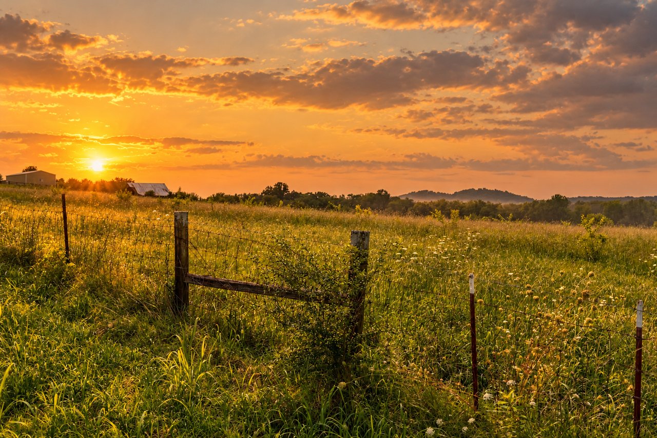 Grassy rural homesite at sunset, with a wire fence, small outbuildings, and wooded hills.