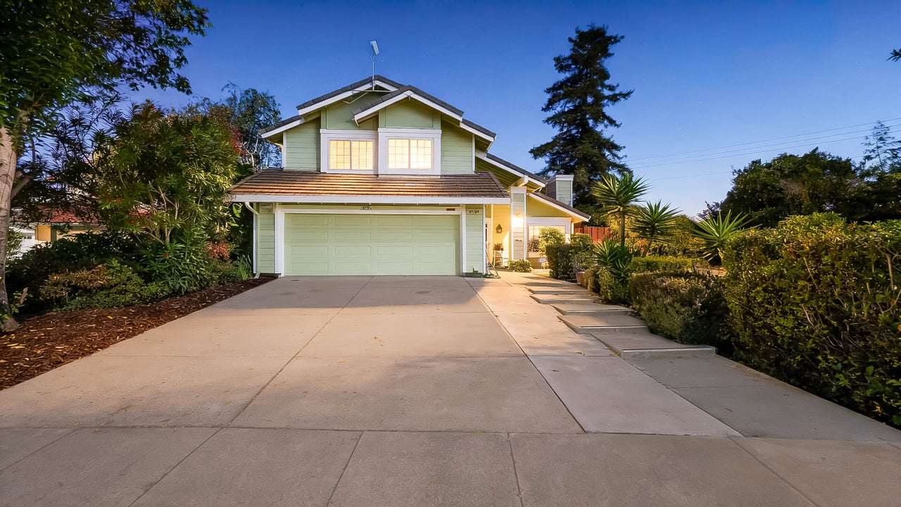 Two-story pale green home with a double garage and broad driveway, warmly lit at dusk.