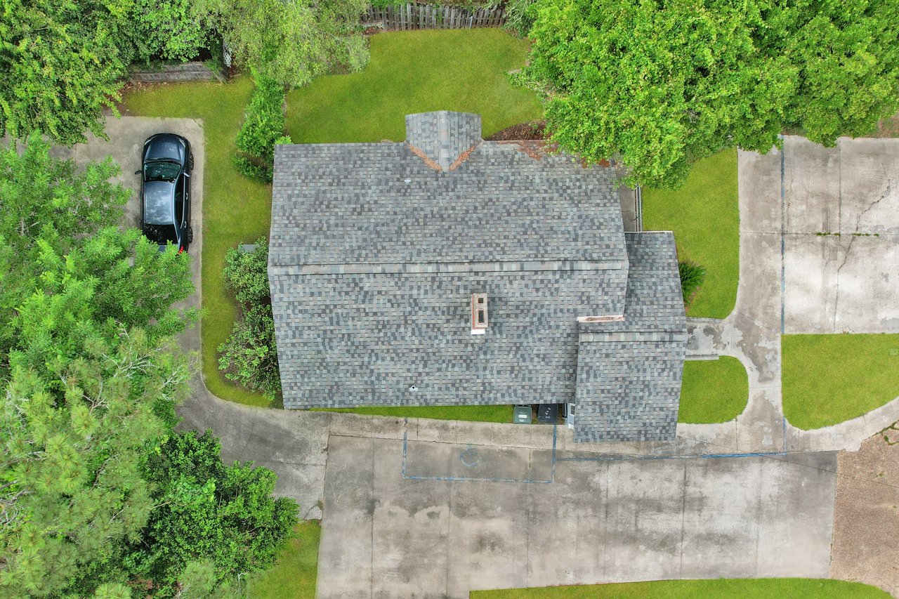 Aerial view of a gray-roofed house surrounded by lush green trees. A black car is parked on a driveway. The scene conveys tranquility.