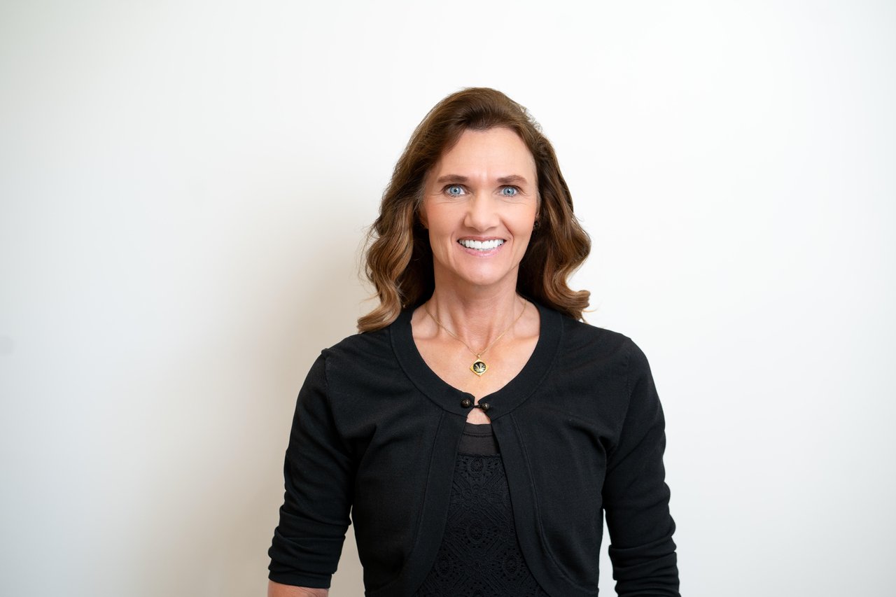 Kelly Little, smiling with long brown hair, wearing a black top and gold pendant necklace.