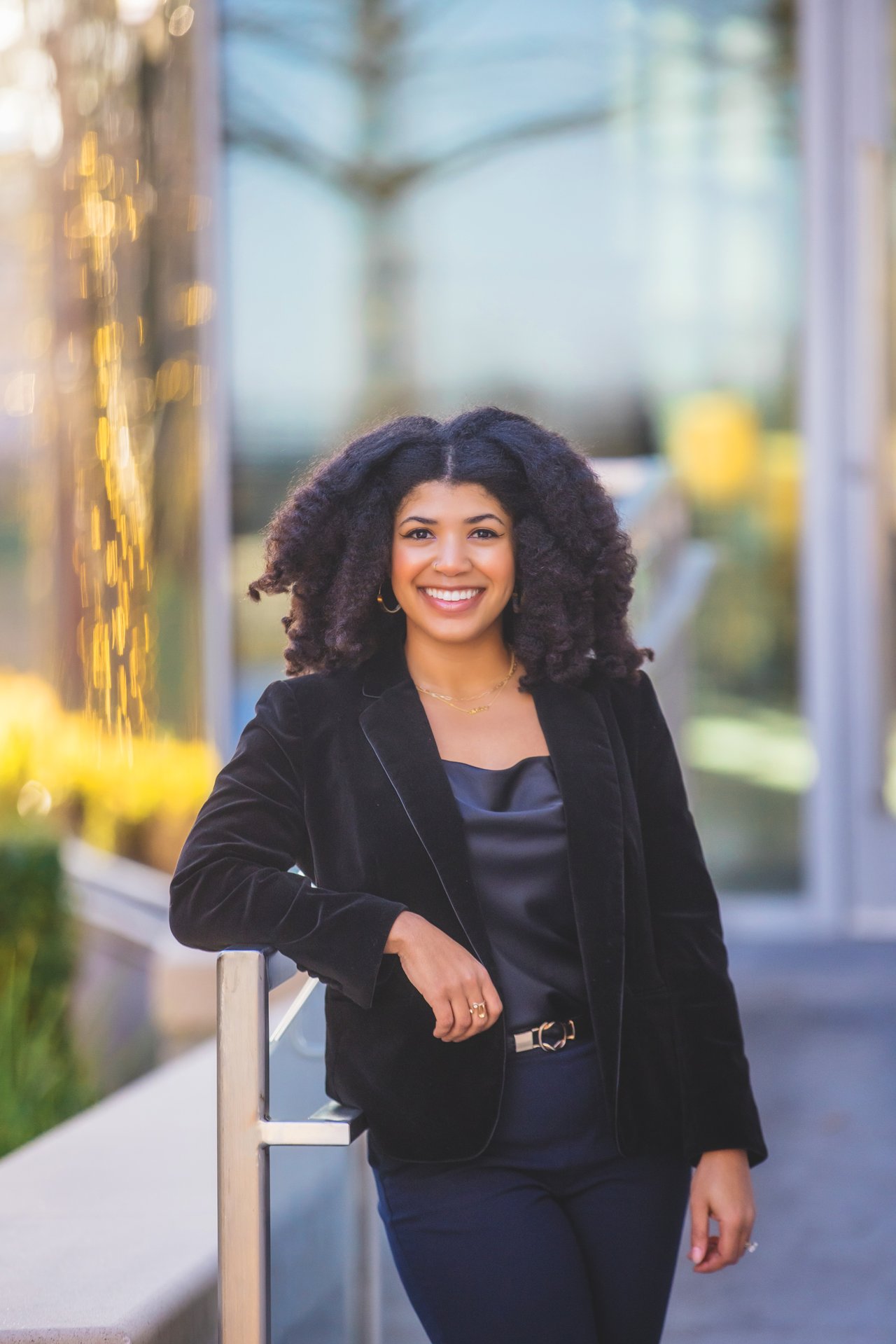 Ashley Junker smiles in a black blazer while leaning on a railing outside a glass-fronted building.