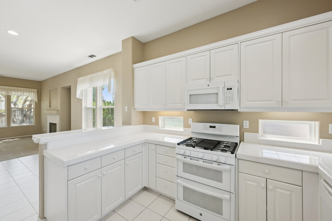 Kitchen with white cabinets, tiled counters, gas range, and a view of the adjoining living area and fireplace.