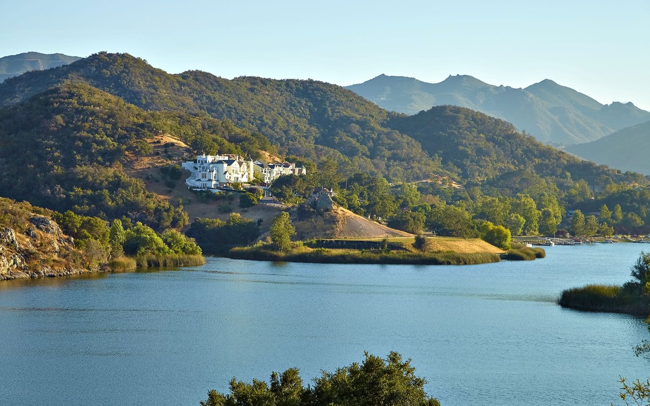White hillside estate overlooking a lake, with wooded mountains in the background.
