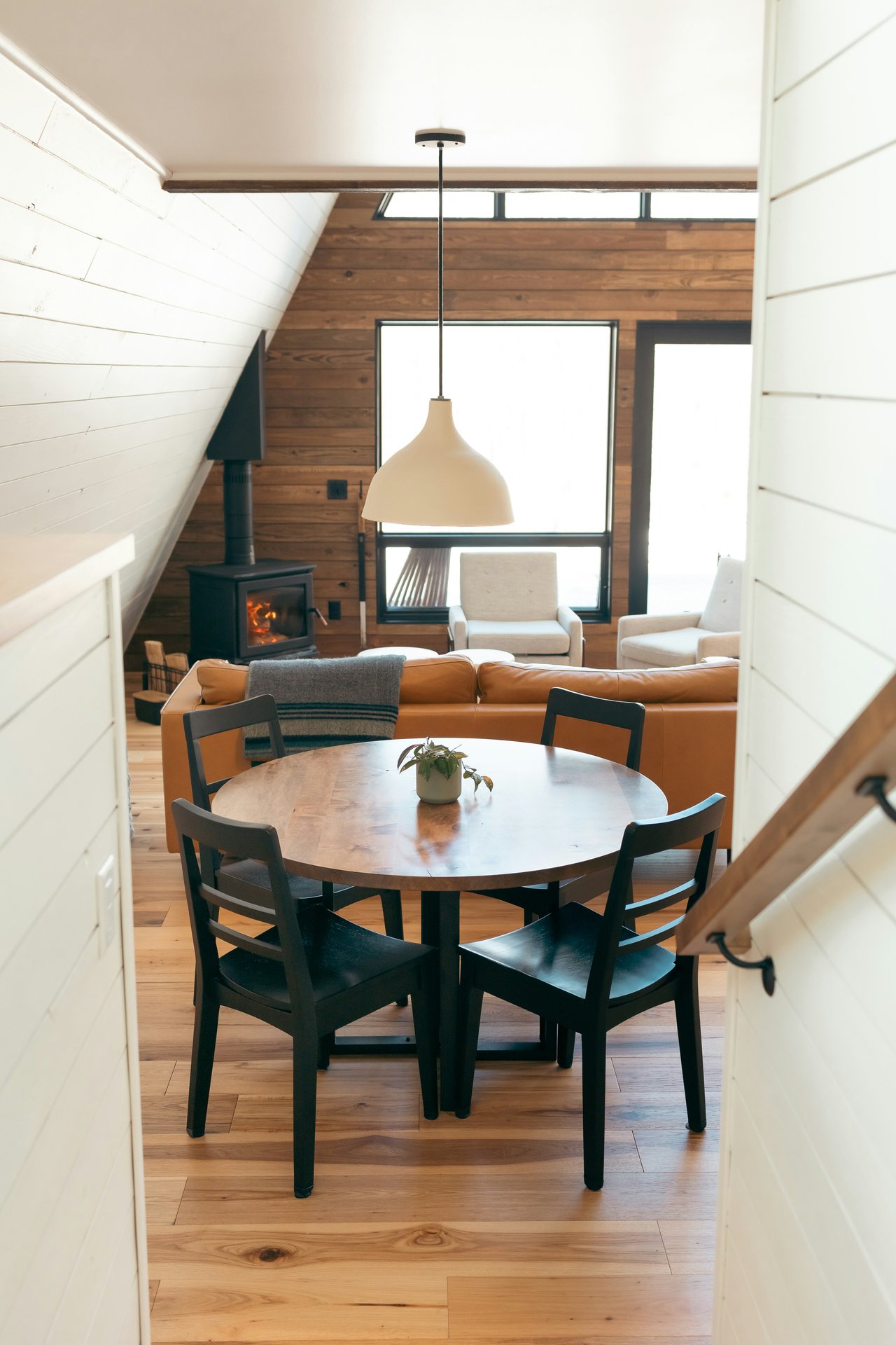 Open-plan dining and living room with a round table, leather sofa, wood stove, and vaulted ceiling.