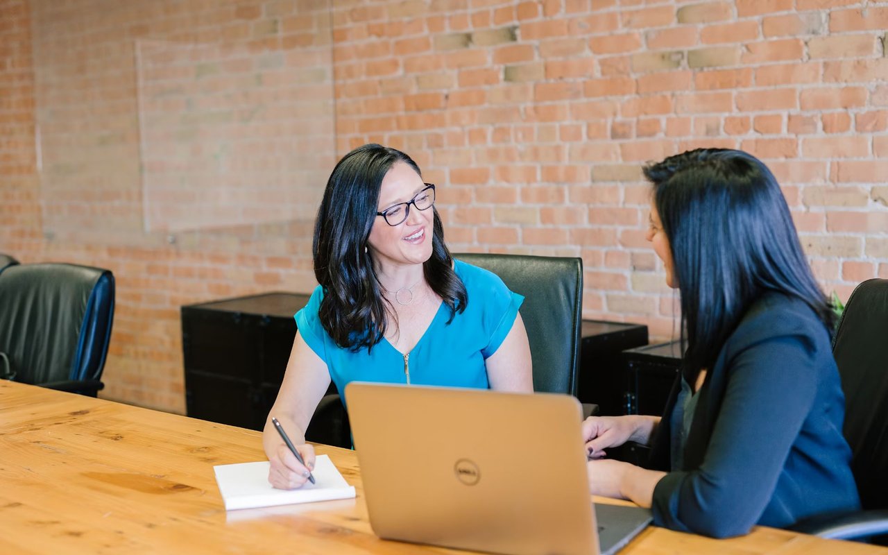 Two women are engaged in a meeting or discussion at a wooden conference table in a room with an exposed brick wall. 