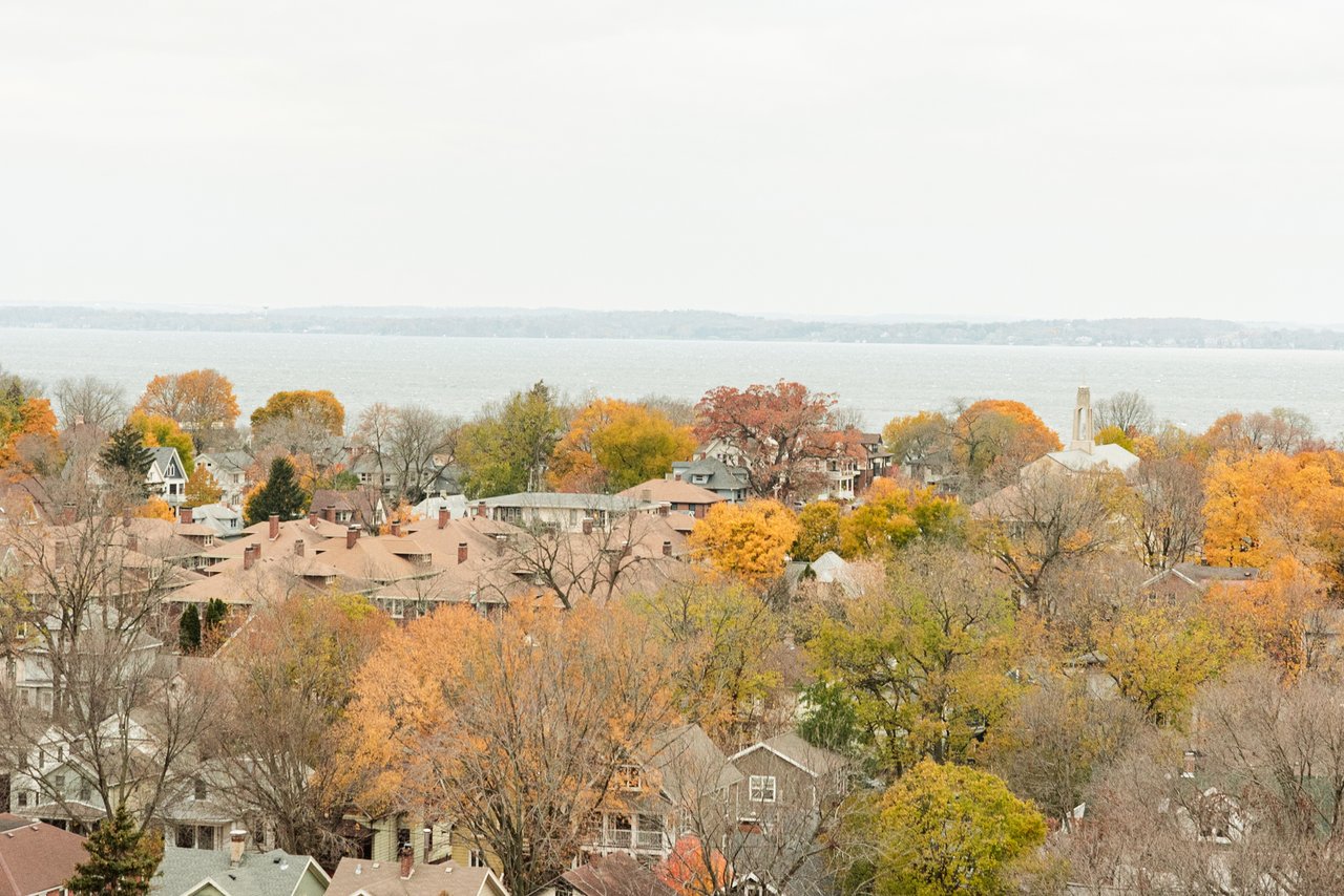 Madison neighborhood rooftops surrounded by yellow and orange fall foliage, with a lake in the background.