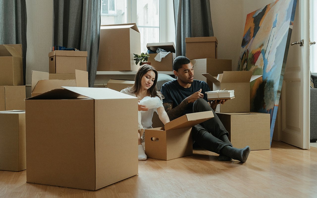 A couple unpacks belongings among moving boxes, with a large colorful artwork leaning nearby.