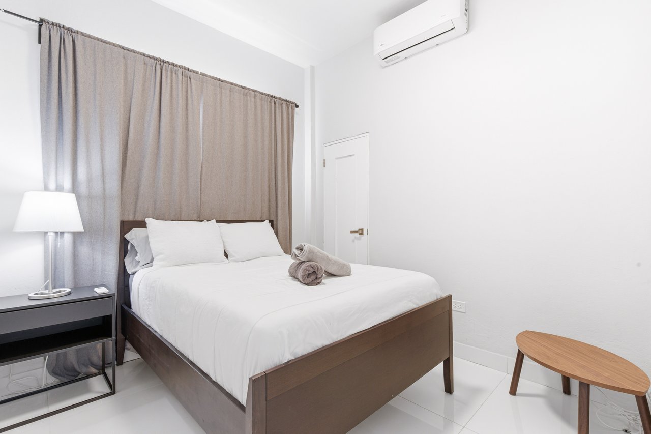 Bedroom with a white bed, gray curtains, bedside lamp, and wall-mounted air conditioner.
