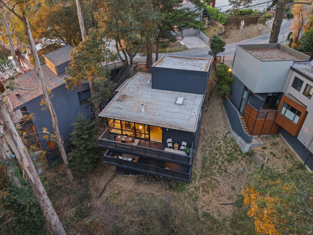Aerial view of a modern dark-sided home with a rooftop deck among autumn trees.