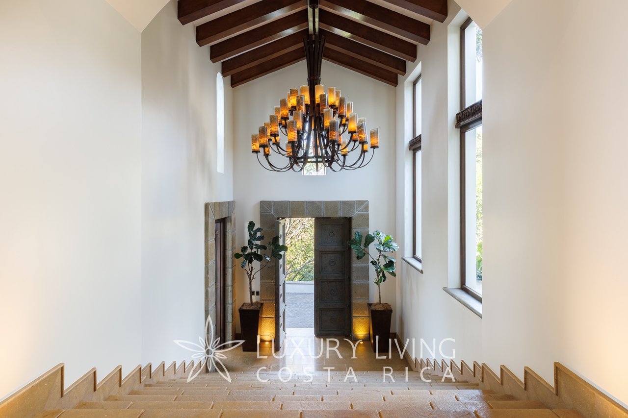 Grand stairway descends toward a stone-framed entry beneath a chandelier and vaulted timber ceiling.