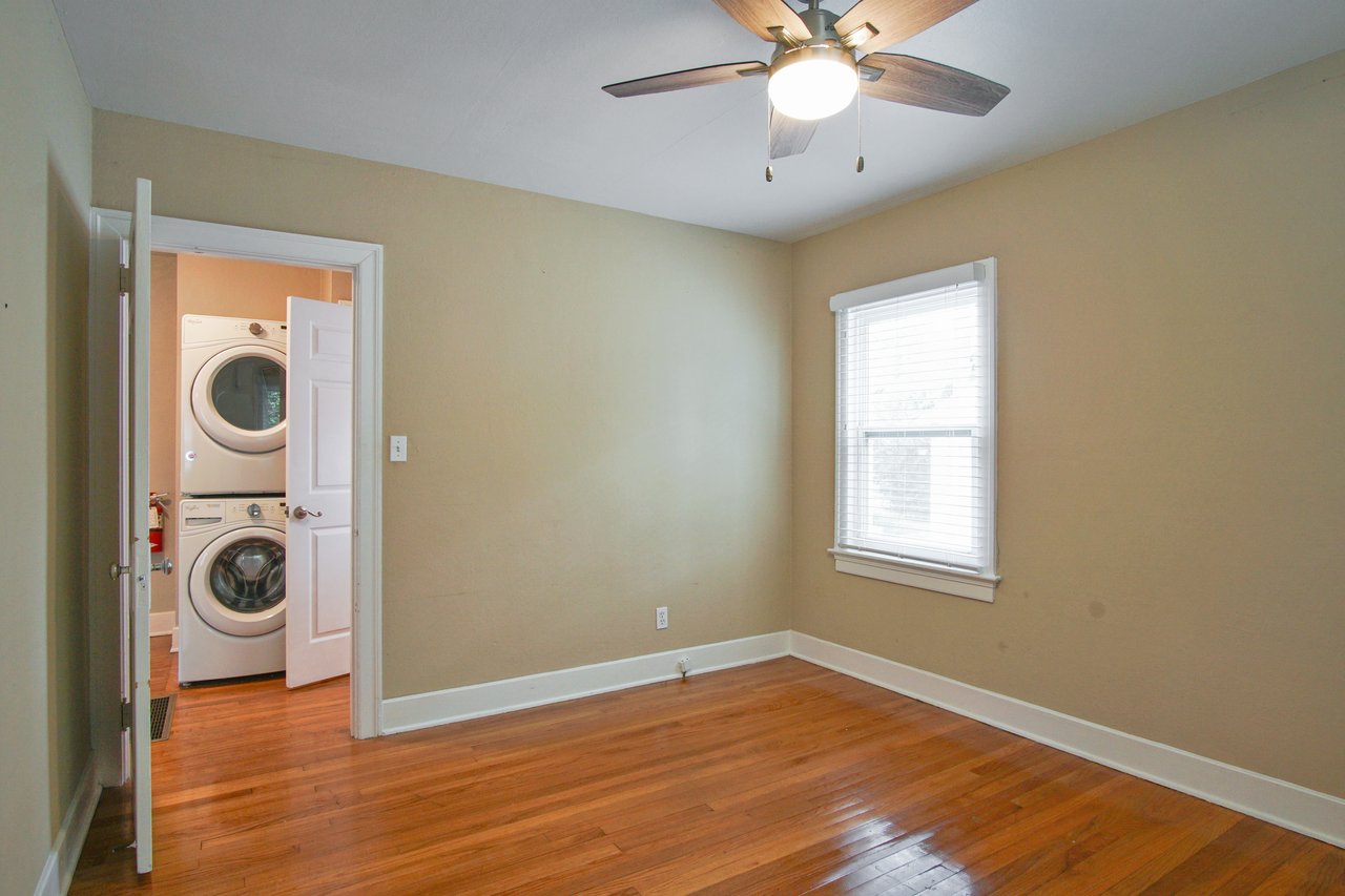 Empty beige room with hardwood floors, a ceiling fan, and a window with blinds. An open door reveals a washer and dryer in a small laundry space.