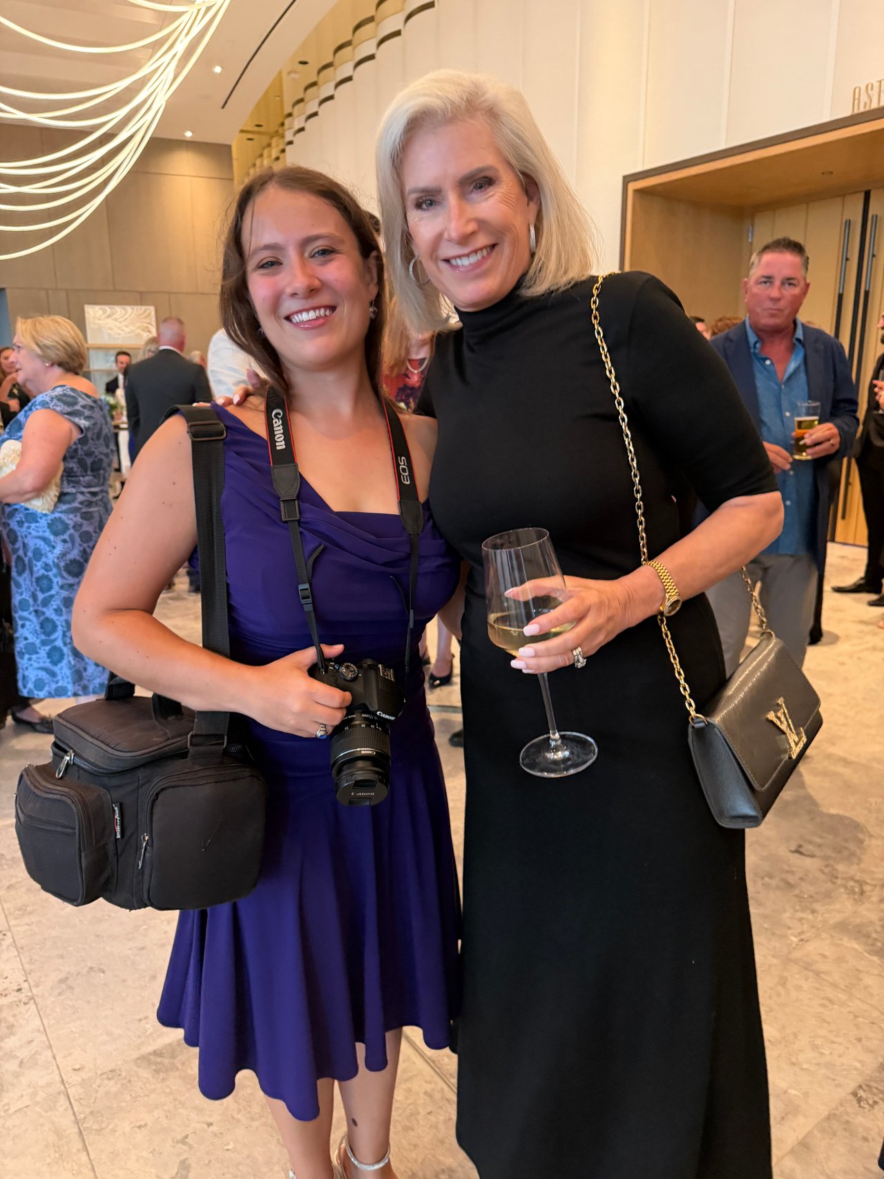 Two women pose at an indoor gathering; one wears a purple dress and camera, the other holds a wine glass.