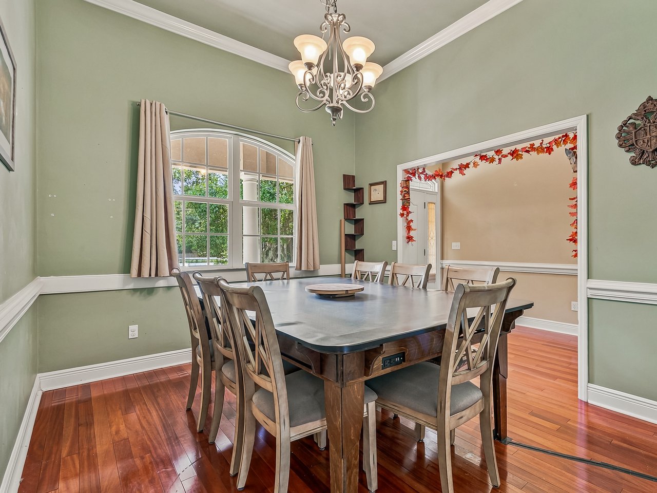 Dining room with a large table beneath a chandelier and a tall arched window.