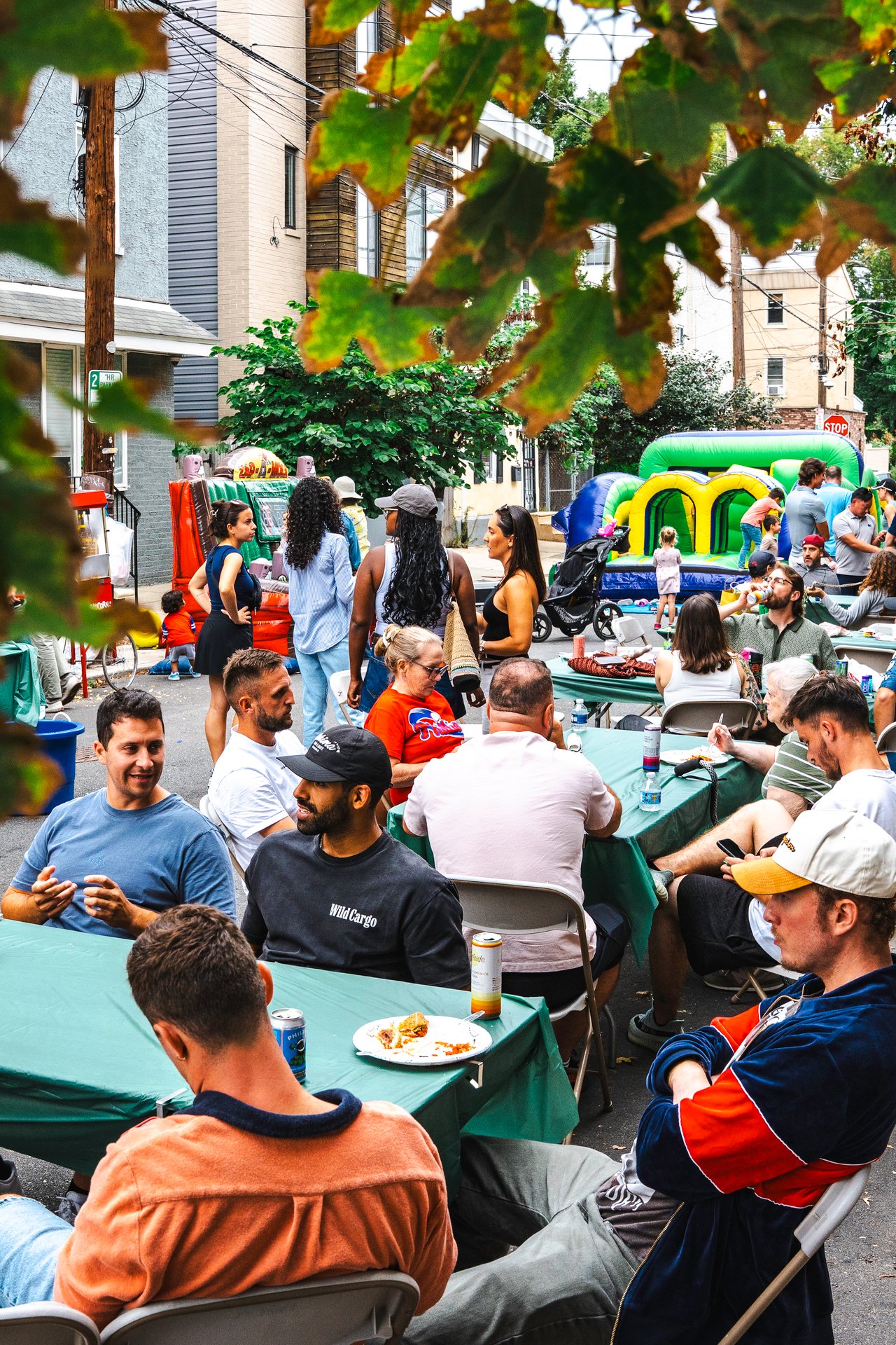 Neighbors eat together at picnic tables during a residential street party, with a bounce house nearby.