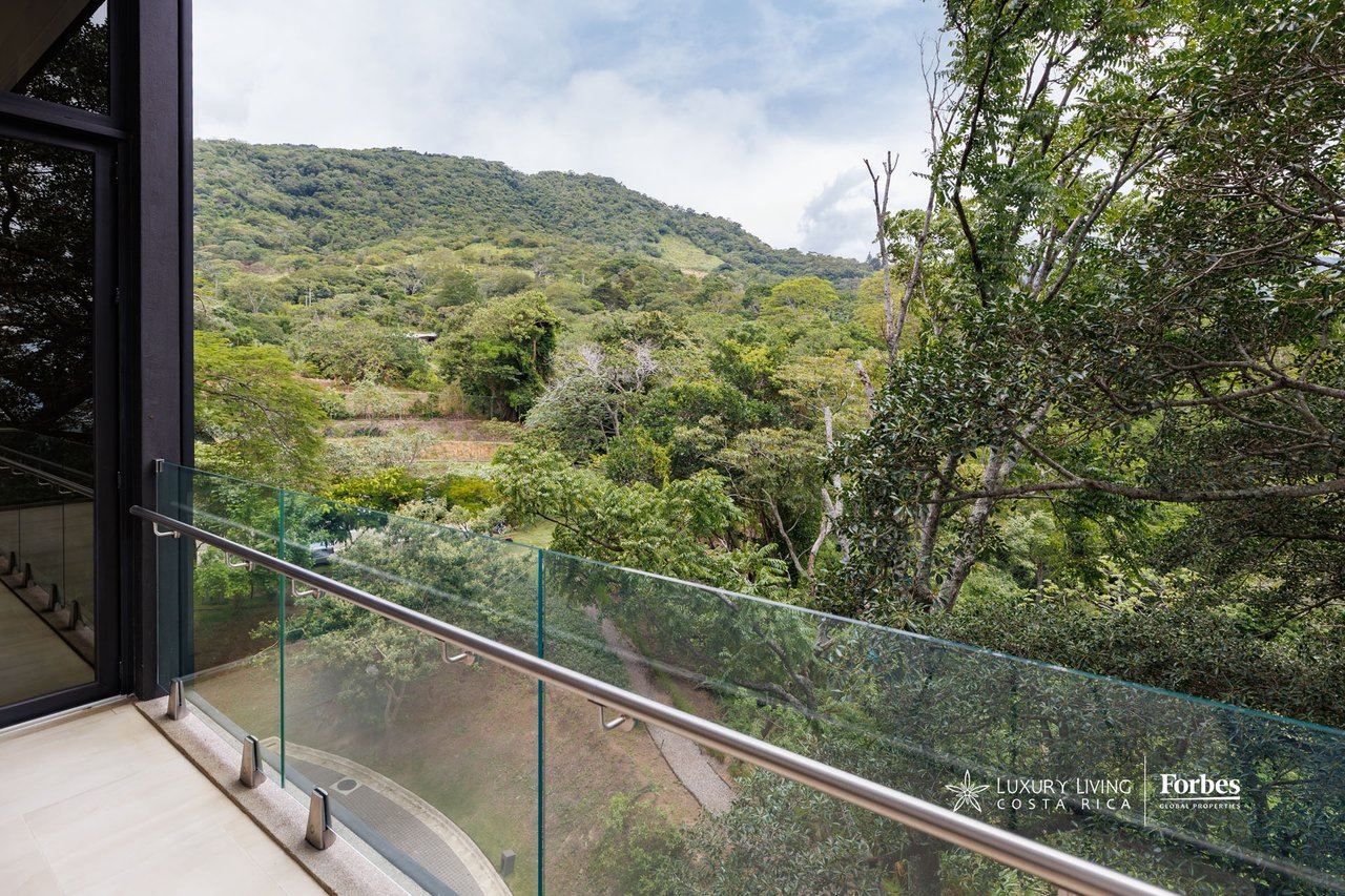 Apartment balcony with glass railing overlooking trees and a green hillside.