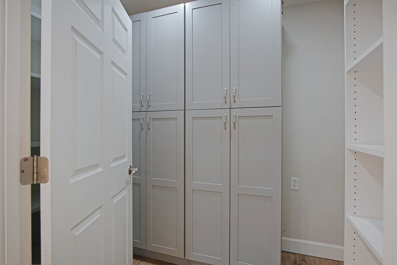 A narrow hallway with closed white cabinets and light wood floor. A slightly open door on the left reveals more shelving, conveying a tidy, organized space.