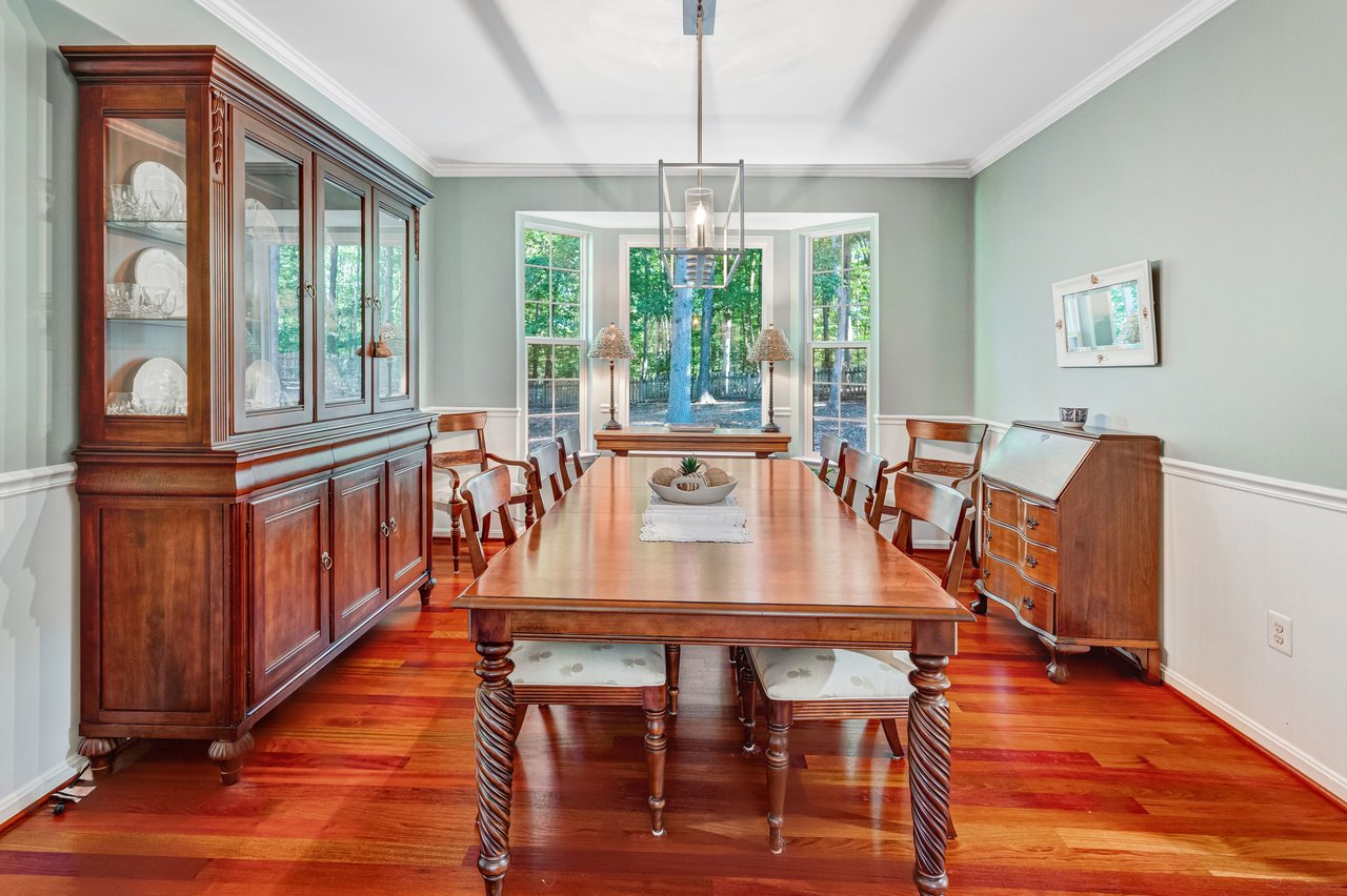 Dining room with a long wooden table, glass-front china cabinet, and windows overlooking trees.